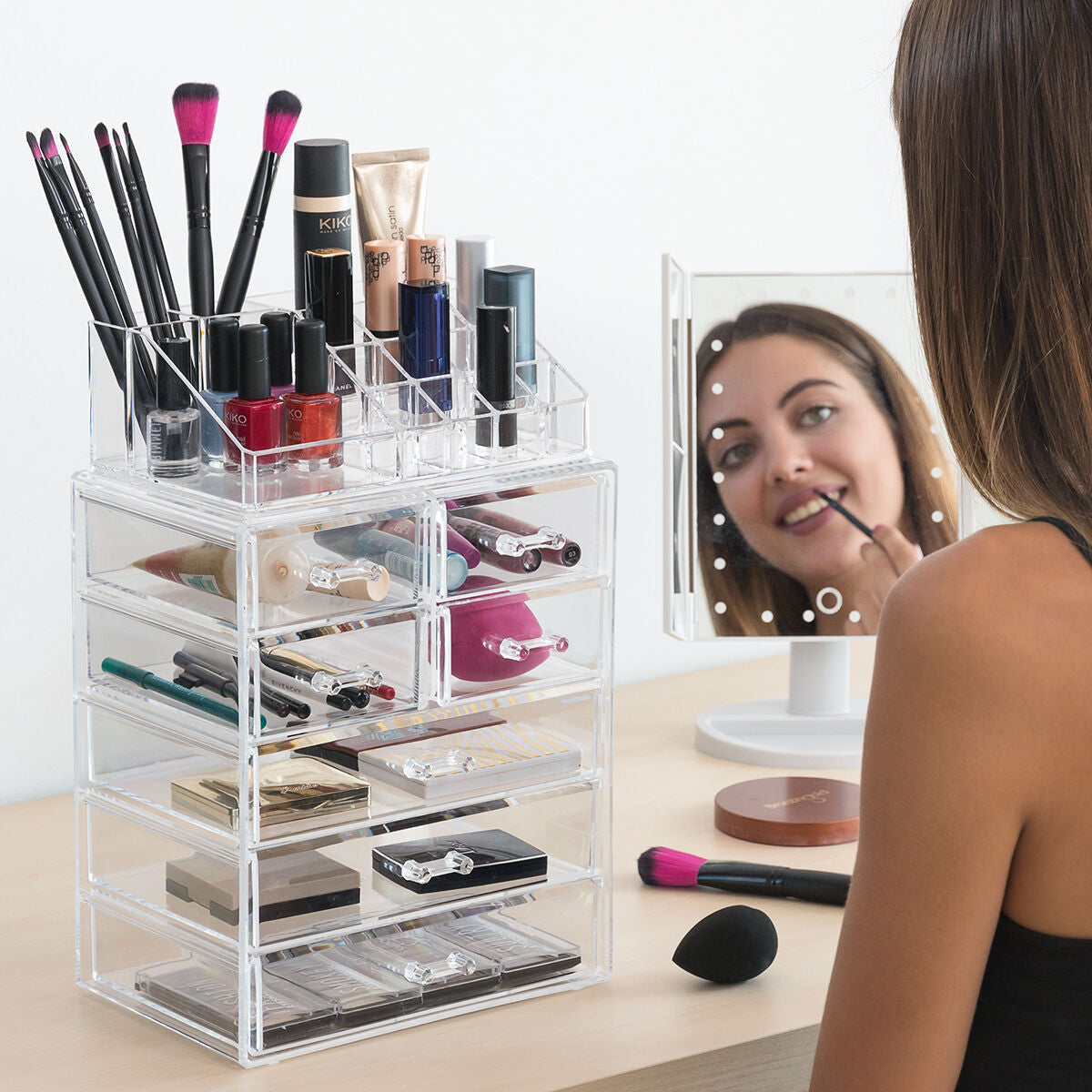Woman using a clear makeup organizer with various cosmetic items on a desk.